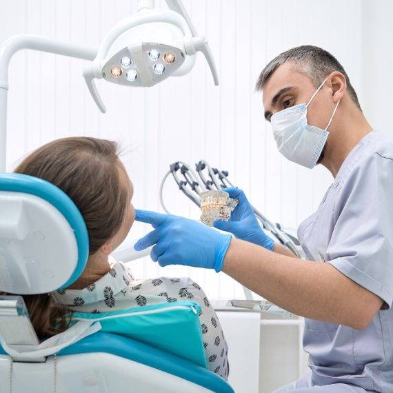 Dentist in mask and gloves shows a dental model to a seated patient in a bright dental clinic during consultation